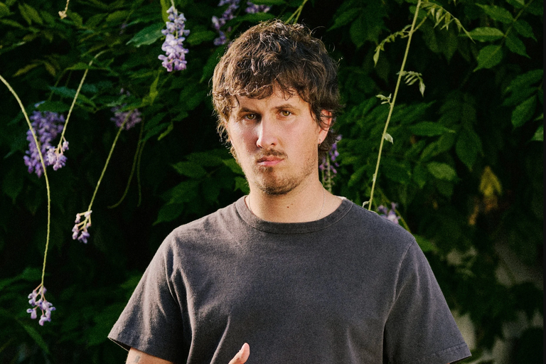 Promotional portrait of musician Arthur Hill wearing a charcoal t-shirt and posing outdoors in front of green leaves and purple wisteria flowers.